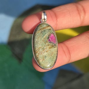 May include: Oval pendant with a silver bail. The pendant features a green stone with a pink inclusion. The stone is set in a silver bezel. The pendant is held by a hand.