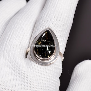 Op de afbeelding: Een zilveren ring met een peervormige, donkergroene edelsteen met gouden en zwarte insluitsels. De edelsteen is gezet in een bezel setting. De ring wordt getoond op een witte ondergrond.