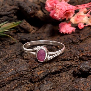 Może przedstawiać: Srebrny pierścionek z żywym owalnym rubinem. Obręcz pierścionka ma podzielony wzór po obu stronach rubinu. Pierścionek jest osadzony na ciemnym, teksturowanym tle z różowymi kwiatami.