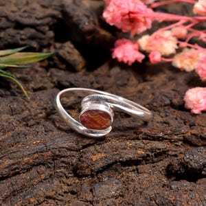 Può includere: Un anello d'argento con una pietra preziosa ovale marrone-rossastra. La fascia dell'anello si curva attorno alla pietra in un design fluido. La gemma ha un aspetto strutturato, su uno sfondo scuro e strutturato.
