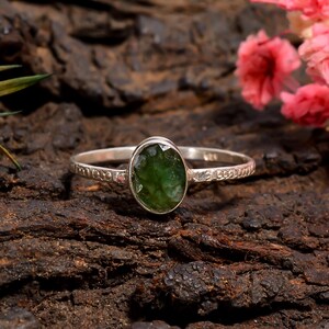 Pode incluir: Um anel de prata esterlina com uma pedra preciosa verde oval. A banda tem um design texturizado. O anel é colocado sobre um fundo escuro e texturizado com flores cor-de-rosa.