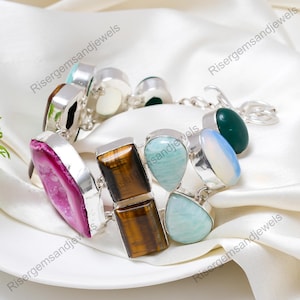 Può includere: Un bracciale in argento con una varietà di pietre preziose, tra cui una grande agata rosa, occhio di tigre e pietre azzurre. Il bracciale è esposto su una superficie bianca e setosa.