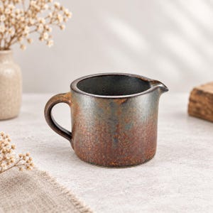May include: A ceramic creamer jug with a dark, mottled finish featuring shades of rust, brown, and charcoal grey. The jug has a rounded body, a small pouring spout, and a curved handle on the side, showcasing a textured, earthy appearance.