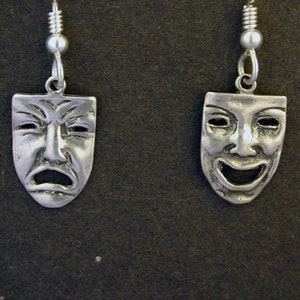 Op de afbeelding: Een paar zilveren oorbellen met een ontwerp van een vrolijk en een verdrietig gezichtsmasker. De maskers zijn gestileerd en hebben een klassieke theatrale uitstraling.