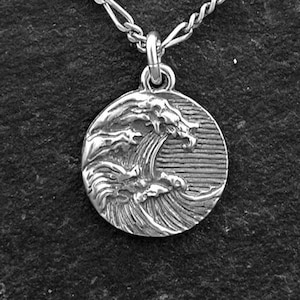 May include: A silver-colored necklace with a round pendant featuring a detailed wave design. The pendant is suspended from a delicate chain. The necklace is displayed on a dark, textured surface.