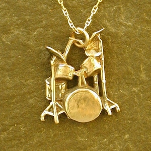 May include: A gold chain necklace with a pendant of a drum set. The pendant is detailed and shows the drums, cymbals, and stands.