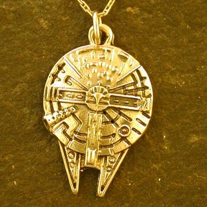 Puede incluir: Un collar con colgante dorado con un diseño detallado del Halcón Milenario. El colgante está suspendido de una delicada cadena dorada. El collar se muestra sobre un fondo texturizado de color verde oscuro.