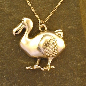 Könnte beinhalten: Eine goldfarbene Halskette mit einem Anhänger in Form eines Dodos. Der Anhänger weist detaillierte Merkmale wie Schnabel, Auge und Flügel auf. Die Halskette ist filigran und goldfarben. Der Anhänger ist etwa 2,5 cm hoch. Der Hintergrund ist dunkel.