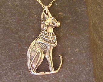Puede incluir: Un collar de oro con un colgante de un gato egipcio estilizado sentado sobre una superficie de piedra gris oscura.