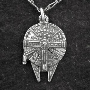 May include: A silver pendant necklace depicting the Millennium Falcon spaceship. The pendant, with its intricate detailing, hangs from a fine chain. The necklace, a nod to the Star Wars series, is set against a dark, textured backdrop.