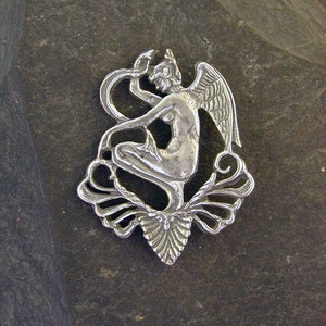 May include: A silver pendant featuring a stylized figure of a woman with wings, holding a snake in her hand. The figure is sitting on a decorative floral design.