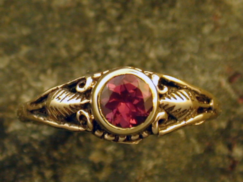 May include: A gold ring with a red gemstone set in a bezel setting. The ring has a delicate floral design on the band.