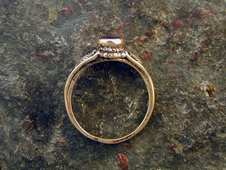 May include: A gold ring with a red gemstone set in a bezel setting. The ring has a simple band and a detailed design around the gemstone.