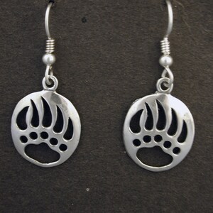 May include: A pair of silver-toned drop earrings featuring a circular pendant with a cut-out bear paw print design. Each earring hangs from a simple silver-toned hook wire with a small decorative bead accent above the circular charm.
