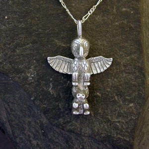 May include: A silver pendant necklace featuring a detailed owl totem with outstretched wings. The owl is perched on top of a smaller figure, possibly a bear or another animal. The necklace is on a silver chain.