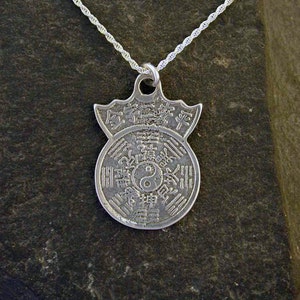 Könnte beinhalten: Silberne Halskette mit einem Anhänger, der ein kreisförmiges Design mit chinesischen Schriftzeichen und einem Yin-Yang-Symbol zeigt. Der Anhänger ist von einem dekorativen Rand umgeben.