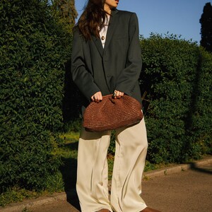 Pu&ograve; includere: Una pochette intrecciata marrone scuro di forma arrotondata &egrave; tenuta davanti a una persona che indossa un blazer verde scuro, un top bianco e pantaloni color crema. La persona indossa scarpe marroni. Lo sfondo comprende cespugli verdi e un cielo blu.