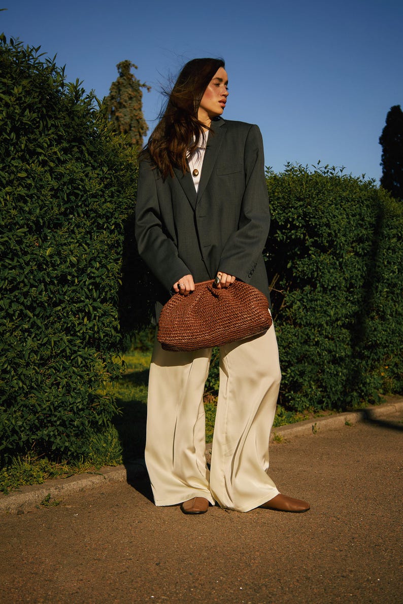 Pu&ograve; includere: Una pochette intrecciata marrone scuro di forma arrotondata &egrave; tenuta davanti a una persona che indossa un blazer verde scuro, un top bianco e pantaloni color crema. La persona indossa scarpe marroni. Lo sfondo comprende cespugli verdi e un cielo blu.