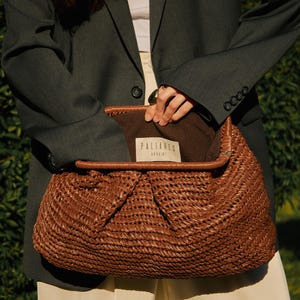 Pu&ograve; includere: Una borsa marrone intrecciata con una parte superiore piegata, che rivela un interno marrone. La borsa ha finiture in pelle e un'etichetta con la scritta "PALIARES UKRAINE". La persona indossa un blazer grigio scuro e pantaloni color crema.