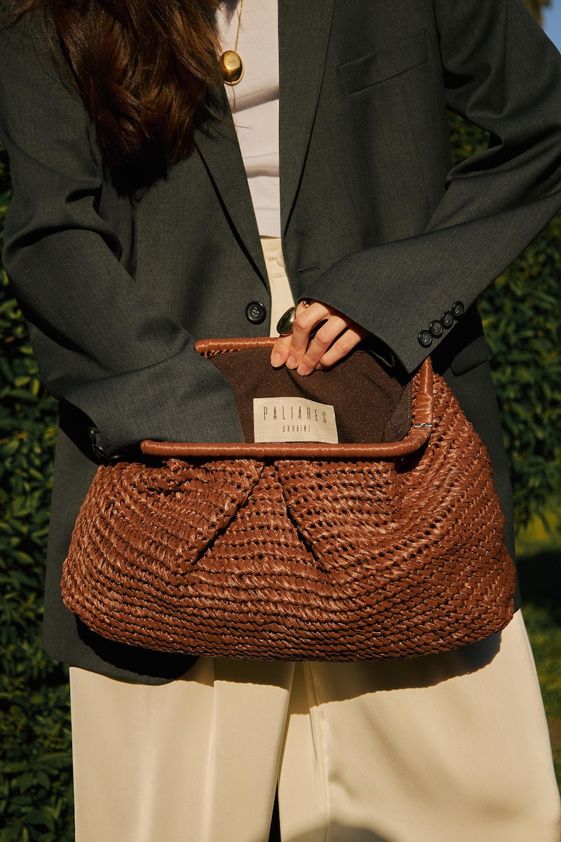 Pu&ograve; includere: Una borsa marrone intrecciata con una parte superiore piegata, che rivela un interno marrone. La borsa ha finiture in pelle e un'etichetta con la scritta "PALIARES UKRAINE". La persona indossa un blazer grigio scuro e pantaloni color crema.