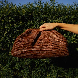 Pu&ograve; includere: Una pochette intrecciata marrone dalla forma arrotondata. La borsa &egrave; tenuta contro uno sfondo di fogliame verde e un cielo azzurro. La pochette ha una superficie strutturata e un design semplice ed elegante.