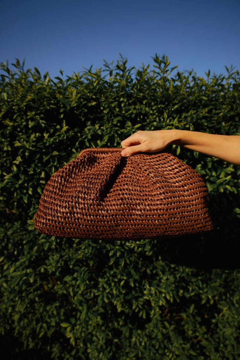 Pu&ograve; includere: Una pochette intrecciata marrone dalla forma arrotondata. La borsa &egrave; tenuta contro uno sfondo di fogliame verde e un cielo azzurro. La pochette ha una superficie strutturata e un design semplice ed elegante.