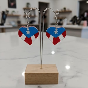 Puede incluir: Pendientes en forma de corazón con el diseño de la bandera de Puerto Rico. Cada pendiente tiene una parte superior azul con una estrella blanca, rayas rojas y blancas y ganchos plateados. Se exhiben en un soporte plateado con una base de madera.