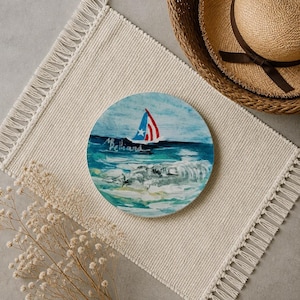 Peut inclure: Un dessous de verre rond en céramique représentant un paysage marin peint avec un voilier arborant un drapeau rouge, blanc et bleu. Le mot Belliard est écrit en lettres blanches sur l'eau bleu foncé. La scène inclut des vagues blanches.