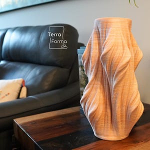 May include: A light brown, textured vase with a draped fabric design. The vase has a cylindrical top and a flowing, sculptural form. The vase is on a wooden table. The logo "Terra Forma" is visible.