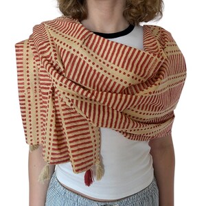 K&ouml;nnte beinhalten: Beige-roter Schal mit einem sich wiederholenden Muster aus vertikalen Streifen und Punkten. Der Schal ist &uuml;ber den Schultern drapiert und hat Quasten an den Enden. Der Schal ist aus einem weichen Stoff gefertigt.