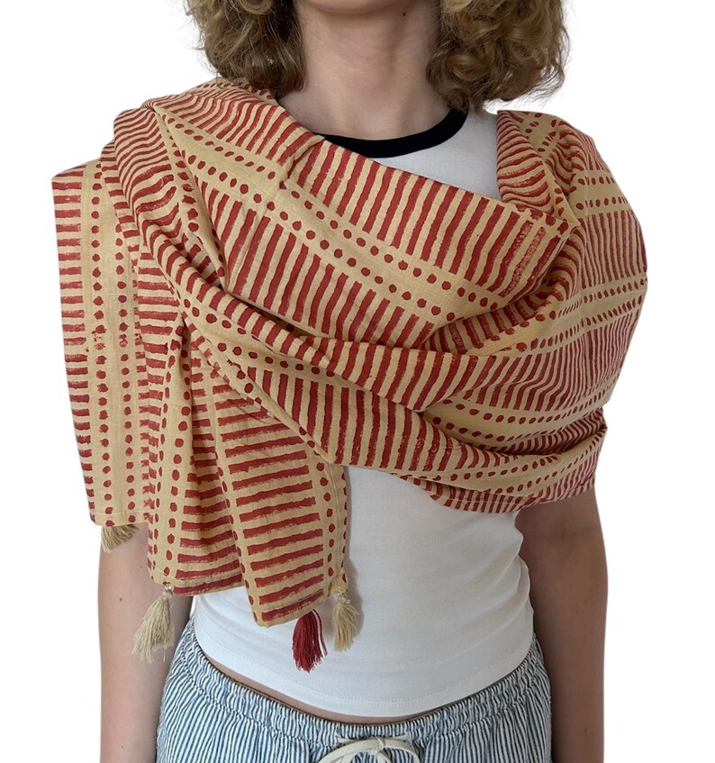 K&ouml;nnte beinhalten: Beige-roter Schal mit einem sich wiederholenden Muster aus vertikalen Streifen und Punkten. Der Schal ist &uuml;ber den Schultern drapiert und hat Quasten an den Enden. Der Schal ist aus einem weichen Stoff gefertigt.