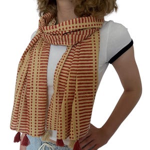 K&ouml;nnte beinhalten: Beige-roter Schal mit Muster und Quasten. Der Schal hat ein sich wiederholendes Muster aus roten Linien und Punkten auf beigem Hintergrund. Die Quasten sind rot und beige. Der Schal ist um den Hals drapiert.
