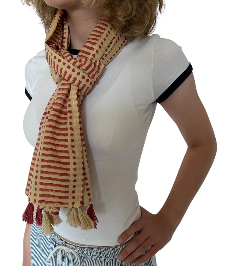 K&ouml;nnte beinhalten: Beige-roter Schal mit Muster und Quasten. Der Schal hat ein sich wiederholendes Muster aus roten Rechtecken und Punkten auf beigem Hintergrund. Die Quasten sind rot und beige.