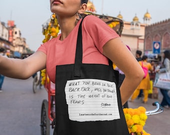 May include: A black tote bag featuring a white rectangular graphic that resembles a piece of lined notebook paper. The text reads: WHAT YOU BRING IN YOUR BACK PACK, WHEN YOU TRAVEL, IS THE WEIGHT OF YOUR FEARS. STEFANO. lastwordsfortheroad.com.