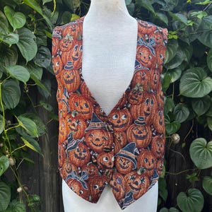 May include: A sleeveless vest featuring an all-over print of orange jack-o'-lanterns with various facial expressions, some wearing black witch hats or blue bows, set against a dark background on a mannequin torso.