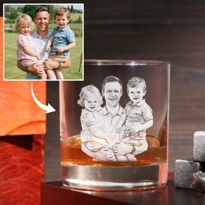 May include: A clear rocks glass with a family portrait etched on the front, filled with amber liquid. The etched image depicts a man holding two children. Beside the glass are three gray whiskey stones and a pair of tongs.