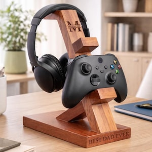 Peut inclure: Support en bois pour casque et manette de jeu avec le texte "BEST DAD EVER". Le support maintient une manette de jeu noire et un casque. Le support est gravé du nom "MALACHI" et du chiffre romain "IV".
