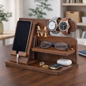 Può includere: Un organizer da scrivania in legno scuro con più scomparti. Presenta un supporto per telefono, porta orologi, un ripiano per occhiali e un piccolo vassoio per accessori. Sono esposti due orologi. L'organizer ha il nome "Malachi" inciso.