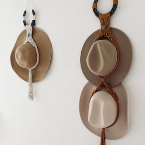 Może przedstawiać: Dwa wieszaki na kapelusze wiszące na białej ścianie. Jeden ma kształt podkowy z turkusowym akcentem, trzymający słomkowy kapelusz. Drugi, również z podkową, trzyma dwa kapelusze, połączone sznurkiem makramy.