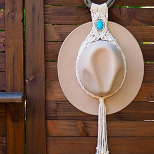 May include: A beige wide-brimmed hat held by a macrame hanger featuring a dark metal horseshoe at the top, cream-coloured cord knots, a central oval turquoise stone in a silver-toned setting, and a long fringe tassel with small wooden beads.