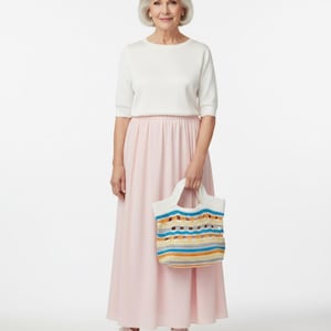 Peut inclure: Une femme porte un haut blanc et une longue jupe rose, tenant un petit sac fourre-tout crocheté coloré. Le sac présente des rayures horizontales bleues, jaunes et beiges. Des baskets blanches complètent la tenue.