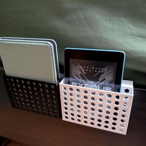 May include: Two bedside organizers, one black and one white, each with a honeycomb pattern. The black organizer holds a light green tablet case, while the white one holds a tablet with a blue frame. The tablet displays text.