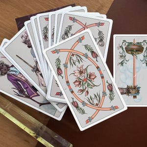 May include: A deck of tarot cards with illustrated backs featuring a central floral design in pink and green on a light grey background. One card shows a figure in a purple coat holding a sword, while another depicts a tall candle with floral accents.
