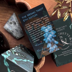 May include: A rectangular card titled The Meraki Tarot 5th Edition with printed text, a signature by Kerri, and a blue floral graphic. The card is positioned near a dark, triangular stone and a portion of a tarot deck box with the text Bouchette & Co.