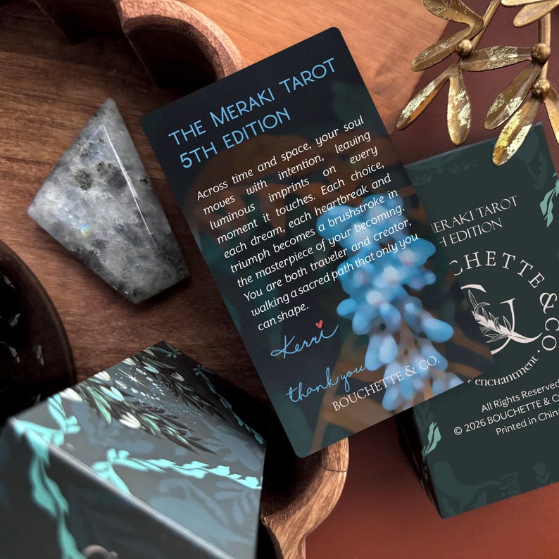 May include: A rectangular card titled The Meraki Tarot 5th Edition with printed text, a signature by Kerri, and a blue floral graphic. The card is positioned near a dark, triangular stone and a portion of a tarot deck box with the text Bouchette & Co.