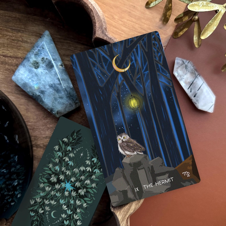 May include: A rectangular tarot card titled The Hermit featuring an owl perched on rocks beneath a crescent moon and a glowing lantern in a dark forest. Beside it lies a second card with a green floral pattern, a blue crystal, and a clear quartz point.