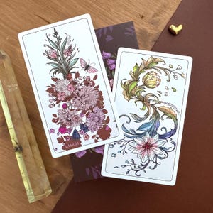 May include: Two rectangular oracle cards with white borders featuring detailed botanical illustrations. One card displays a bouquet of pink and brown flowers with a small butterfly and the word surrender. The other shows swirling floral vines and the word contraction.