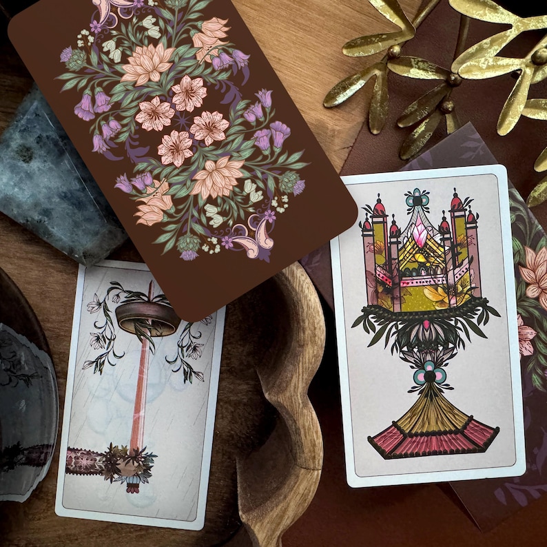 May include: Three rectangular tarot cards with illustrated backs and fronts. One card back features a dark brown background with pink and purple floral patterns. The card fronts display detailed line art of a candle and a decorative chalice.
