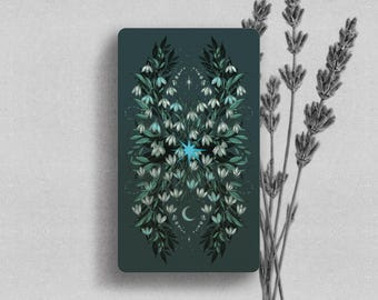 May include: A rectangular tarot card with a dark teal background featuring a symmetrical illustration of white snowdrop flowers and green leaves. A bright blue starburst sits in the center, with a small crescent moon below and a tiny star above.