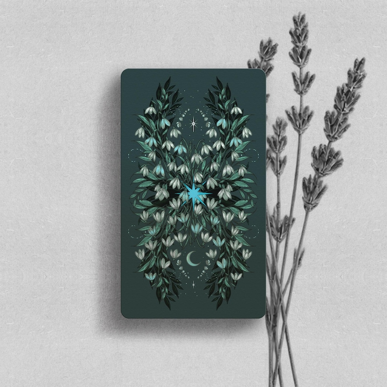 May include: A rectangular tarot card with a dark teal background featuring a symmetrical illustration of white snowdrop flowers and green leaves. A bright blue starburst sits in the center, with a small crescent moon below and a tiny star above.